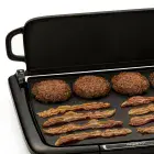 A close-up of the Cool-touch Electric Griddle/Warmer Plus cooking bacon and hamburgers with the tray up to act as a spatter shield.