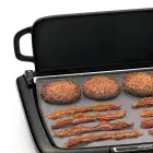 A close-up of the Cool-touch Electric Griddle/Warmer Plus cooking bacon and hamburgers with the tray up to act as a spatter shield.