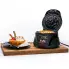 The Belgian Waffle Bowl Maker on a cutting board with the cover open and a waffle bowl on the cooking grid. A filled waffle bowl is on a plate to the left of the unit.